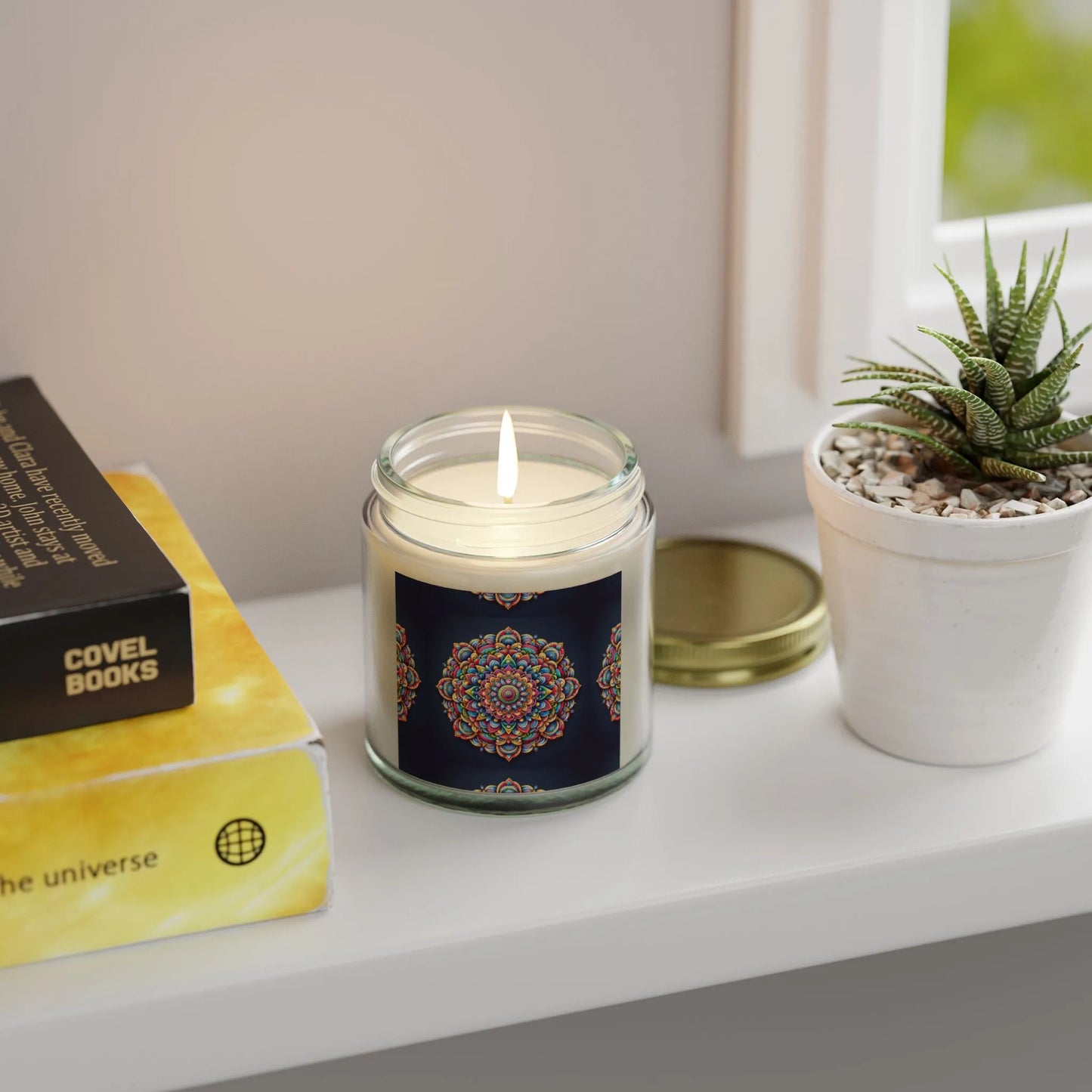 A Zen Candle from Printify, crafted with eco-friendly coconut apricot wax and featuring a vibrant mandala design, sits on a windowsill. Nearby, a potted succulent complements the stacked books highlighting "COVEL BOOKS." The soft light streaming through the window creates an ideal atmosphere for relaxation or meditation.