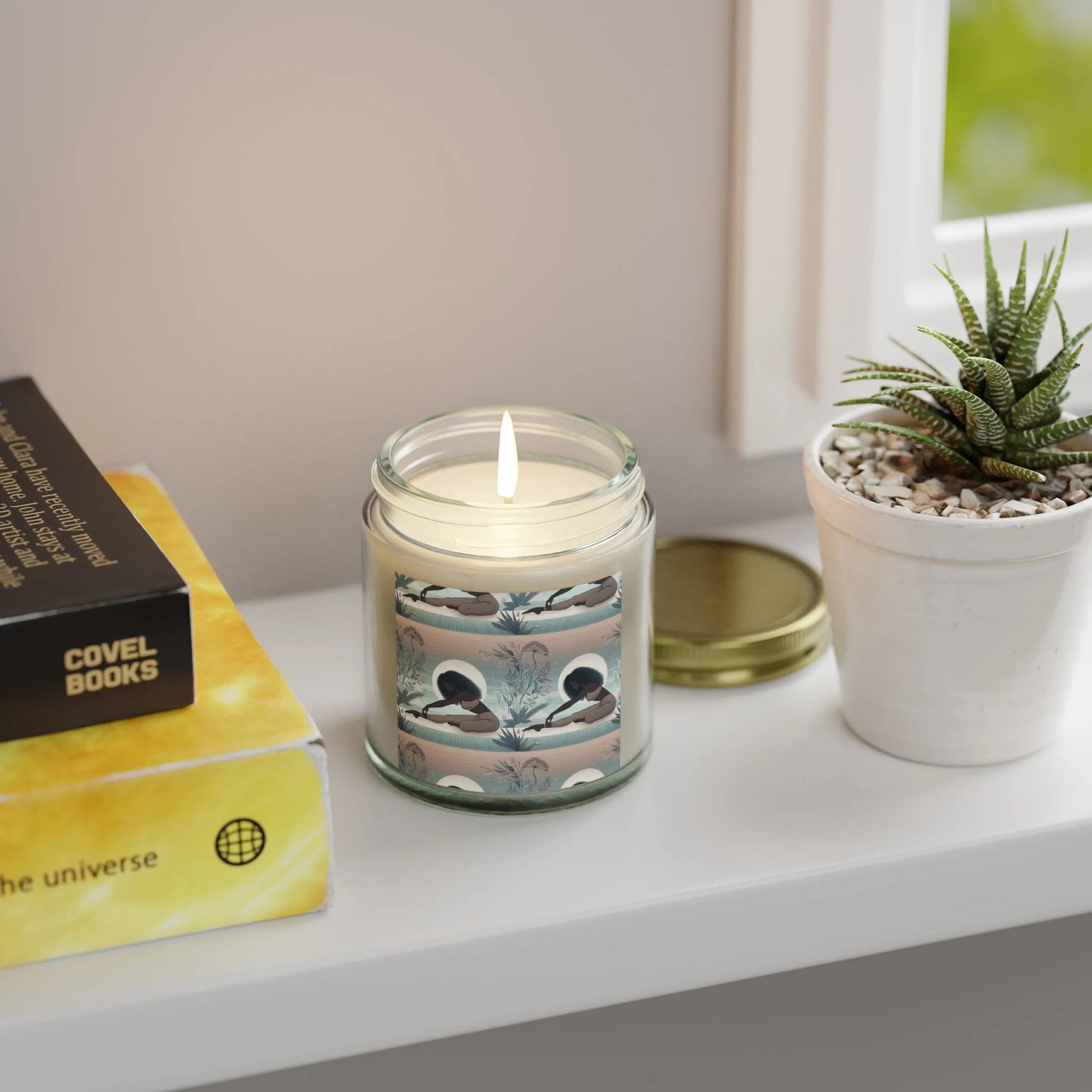 On a windowsill sits a lit Yoga Girl Aroma Scented Candle by Printify, crafted from eco-friendly Coconut Apricot Wax and encased in a glass jar adorned with a celestial-themed label. Next to it, there's a small potted succulent and a stack of books, featuring one with a yellow cover and circular design.