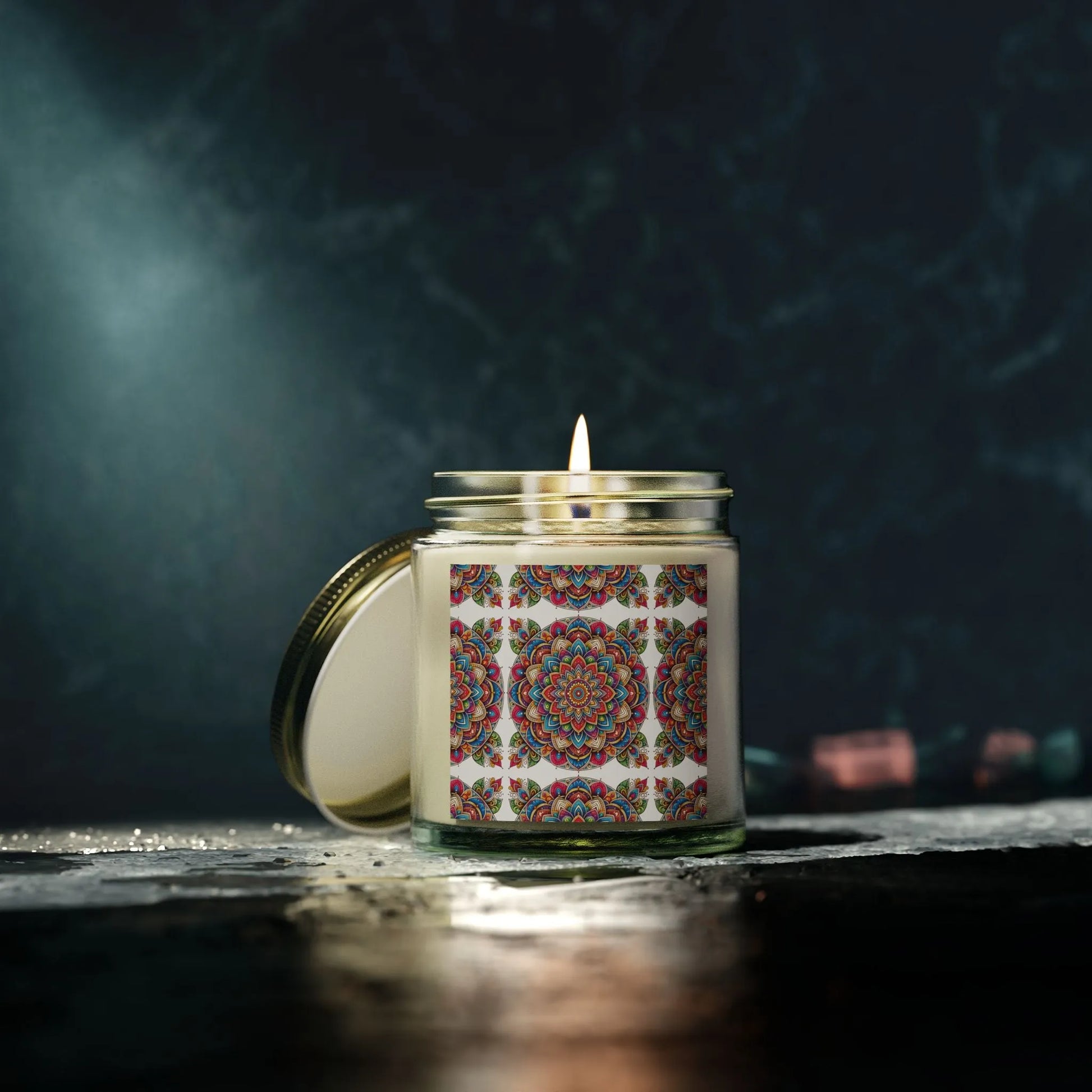 An Aromatherapy Candle Essential Oil by Printify, featuring a vibrant mandala design on its glass jar, illuminates the setting. Crafted from eco-friendly wax, it rests on a textured surface under gentle lighting, creating a cozy atmosphere against the dark backdrop—ideal for yoga or zen relaxation.