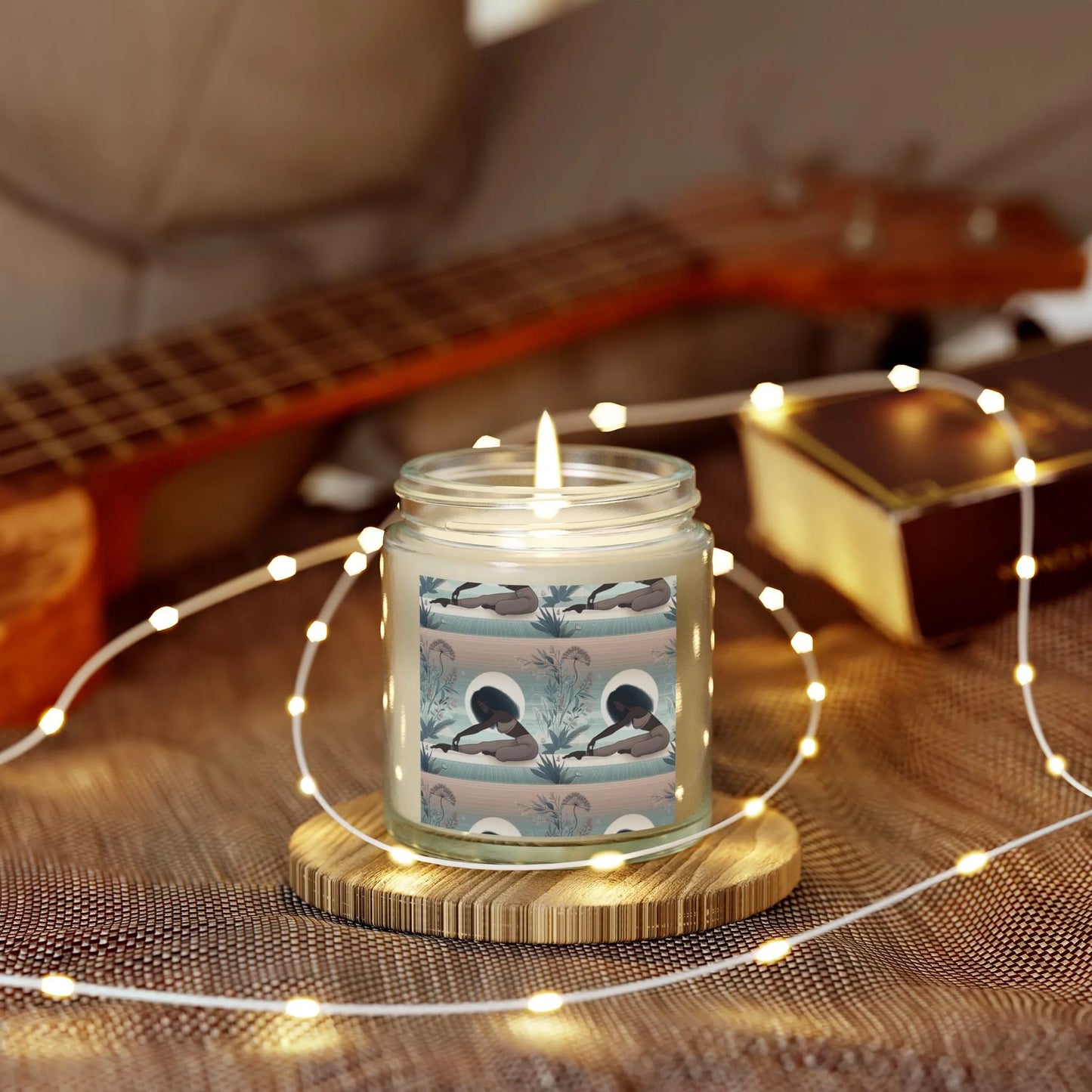 The Yoga Girl Aroma Scented Candle by Printify, set in a glass jar with artistic labels and crafted from eco-friendly wax, rests on a wooden coaster. Warm fairy lights encircle it as a ukulele and a closed book are partially visible on the textured surface behind.