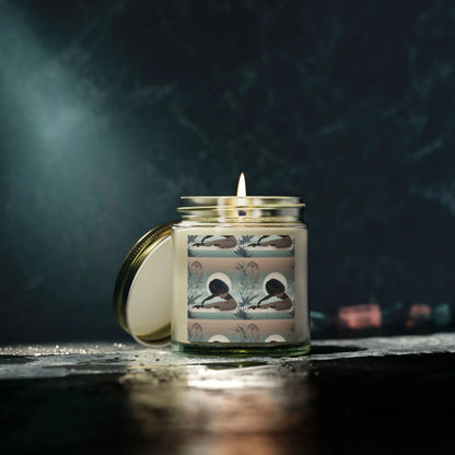 The Yoga Girl Aroma Scented Candle by Printify features eco-friendly wax in a glass jar, adorned with a floral label of a person lying among plants. This Zen candle rests on a dark, reflective surface with a softly lit and shadowed background.