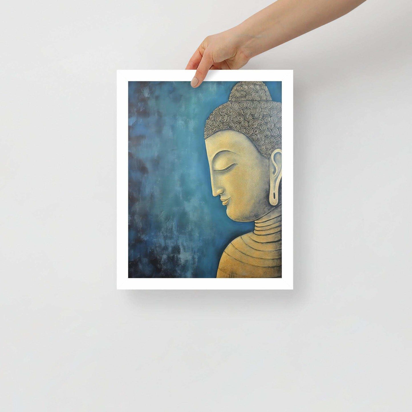 A hand is placing a framed poster on a white background, where a serene Golden Buddha Art with a patterned head and golden features is portrayed in profile against a mottled blue backdrop, all within a white oak frame.