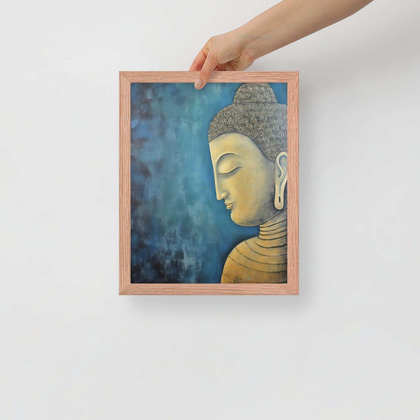 A hand is placing a framed poster on a white background, where a serene Golden Buddha Art with a patterned head and golden features is portrayed in profile against a mottled blue backdrop, all within a red oak frame.