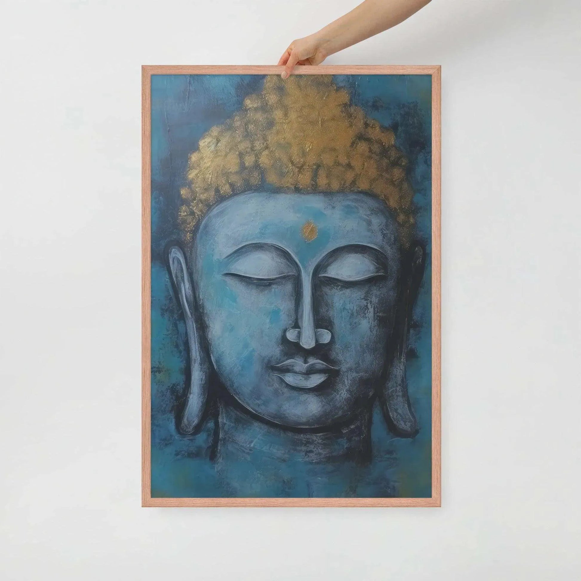 A hand is adjusting a framed poster with a red oak finish depicting a blue Buddha head with a textured gold leaf crown against a blue background, giving the impression of tranquility and artistic simplicity against a white backdrop.