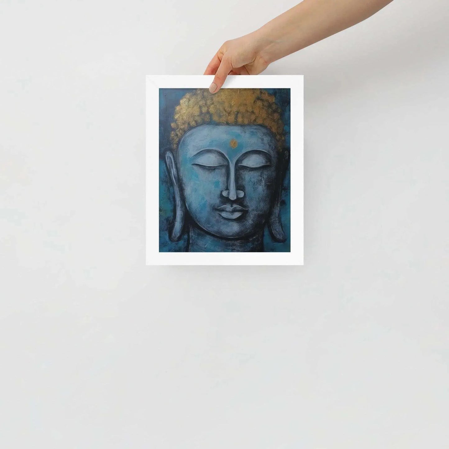 A hand is adjusting a framed poster with a white oak finish depicting a blue Buddha head with a textured gold leaf crown against a blue background, giving the impression of tranquility and artistic simplicity against a white backdrop.