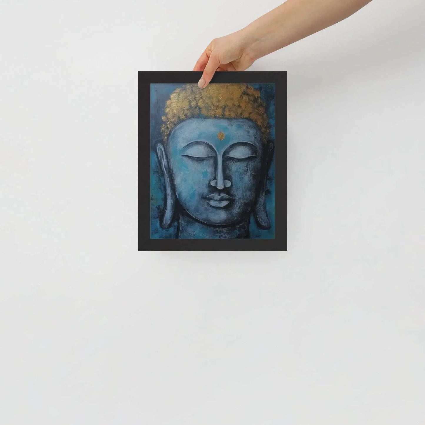 A hand is adjusting a framed poster with a black oak finish depicting a blue Buddha head with a textured gold leaf crown against a blue background, giving the impression of tranquility and artistic simplicity against a white backdrop.