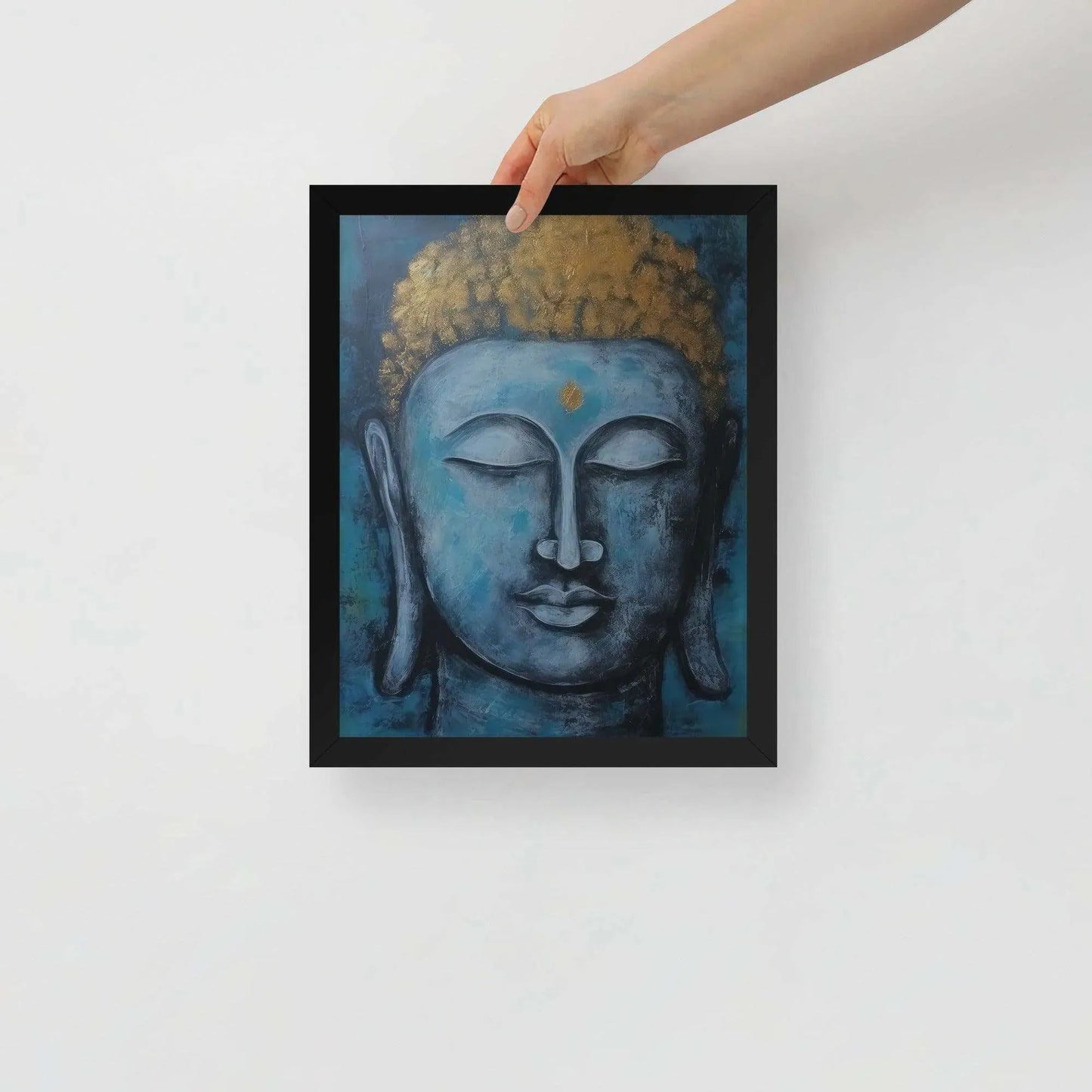 A hand is adjusting a framed poster with a black oak finish depicting a blue Buddha head with a textured gold leaf crown against a blue background, giving the impression of tranquility and artistic simplicity against a white backdrop.