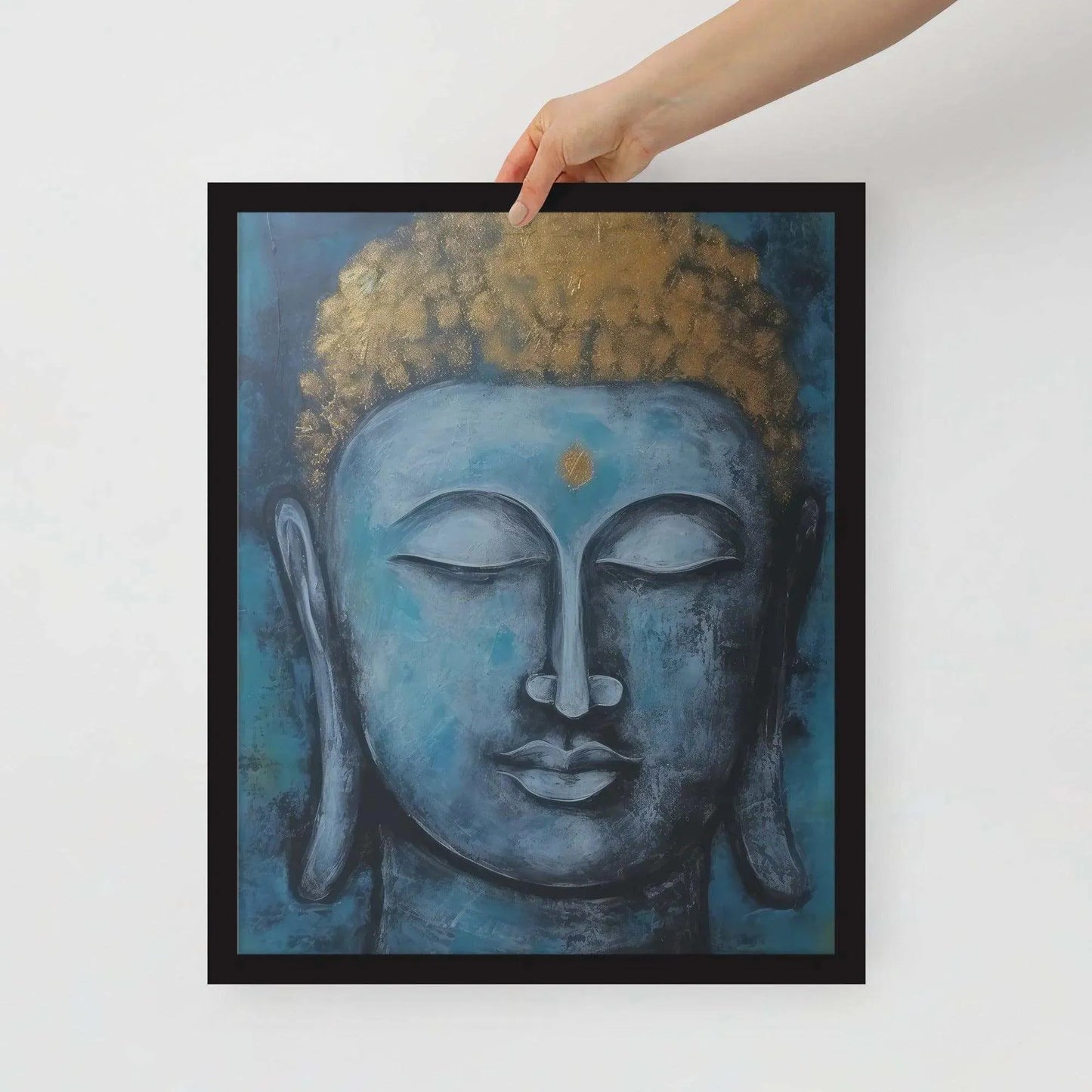 A hand is adjusting a framed poster with a black oak finish depicting a blue Buddha head with a textured gold leaf crown against a blue background, giving the impression of tranquility and artistic simplicity against a white backdrop.