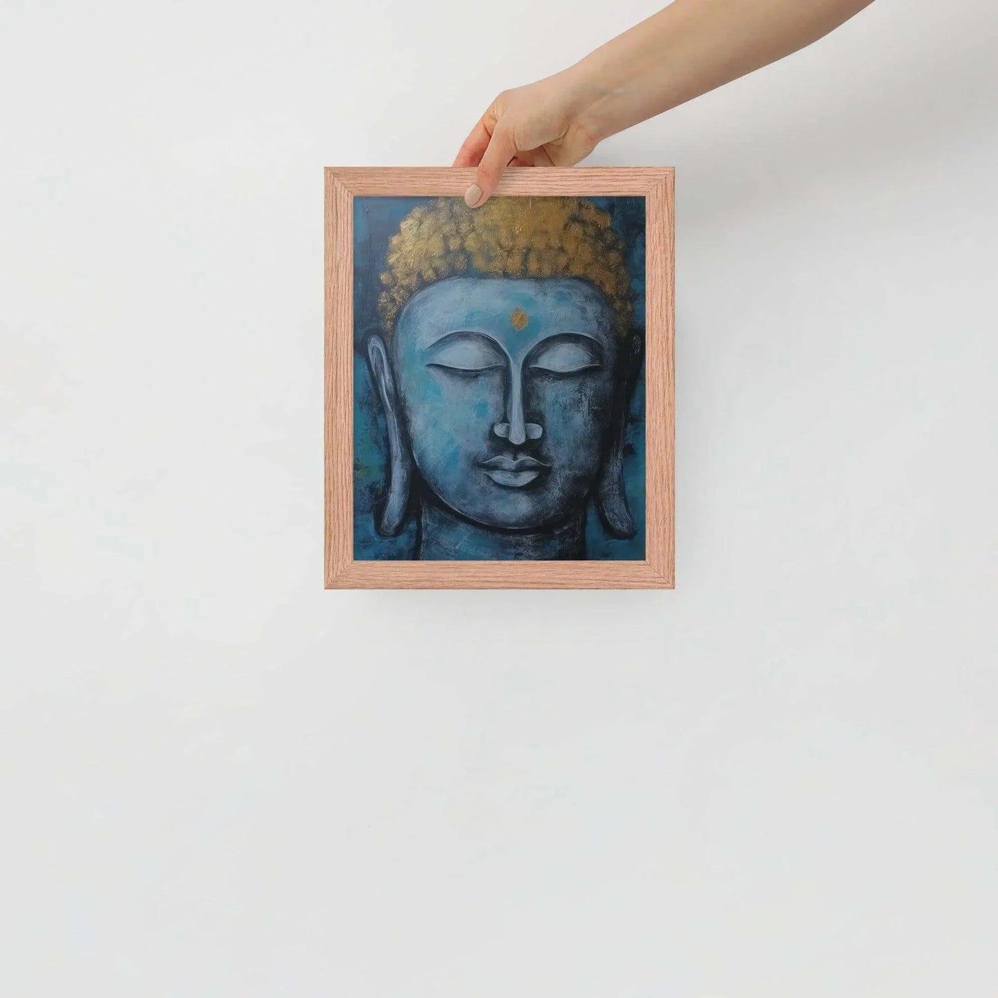 A hand is adjusting a framed poster with a red oak finish depicting a blue Buddha head with a textured gold leaf crown against a blue background, giving the impression of tranquility and artistic simplicity against a white backdrop.