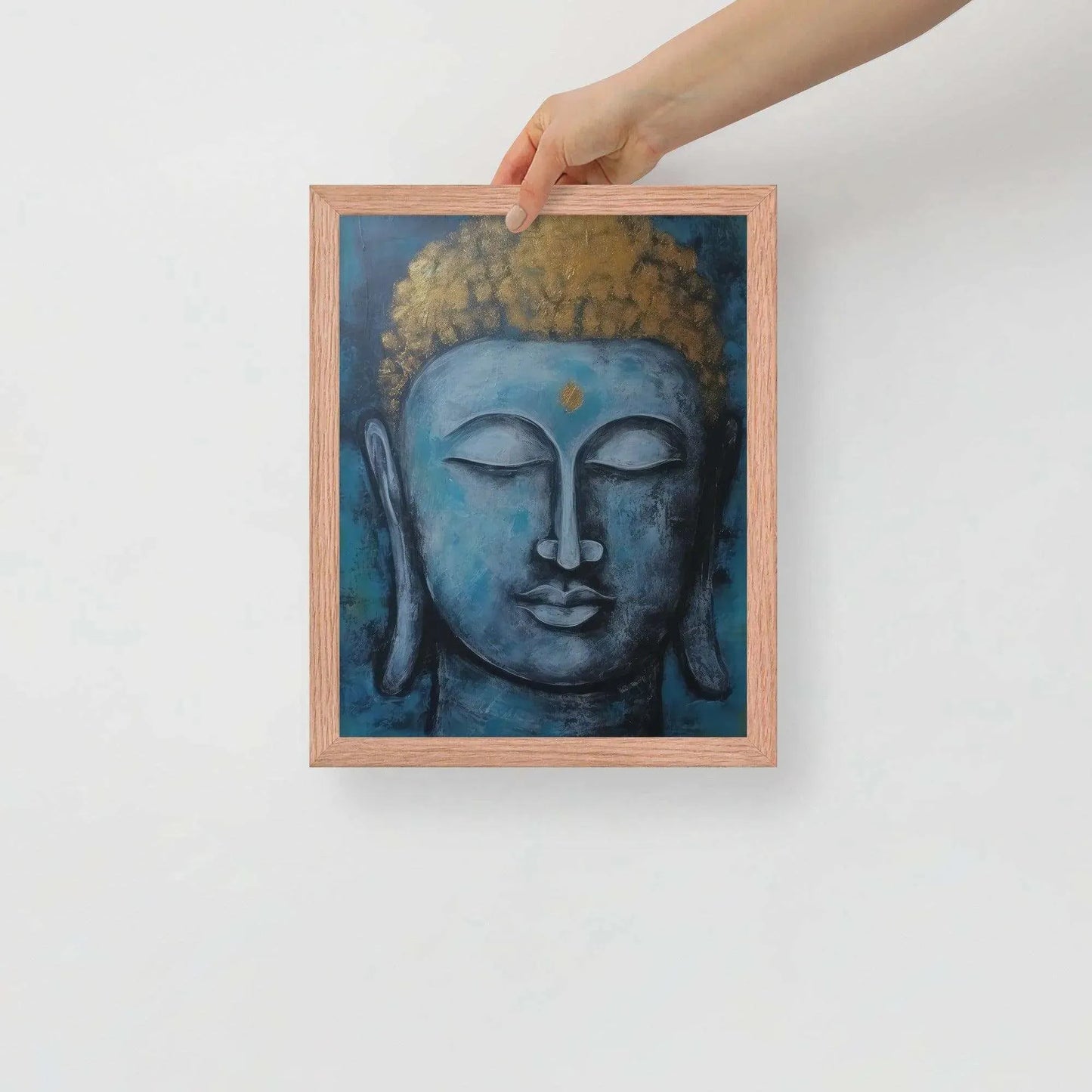 A hand is adjusting a framed poster with a red oak finish depicting a blue Buddha head with a textured gold leaf crown against a blue background, giving the impression of tranquility and artistic simplicity against a white backdrop.