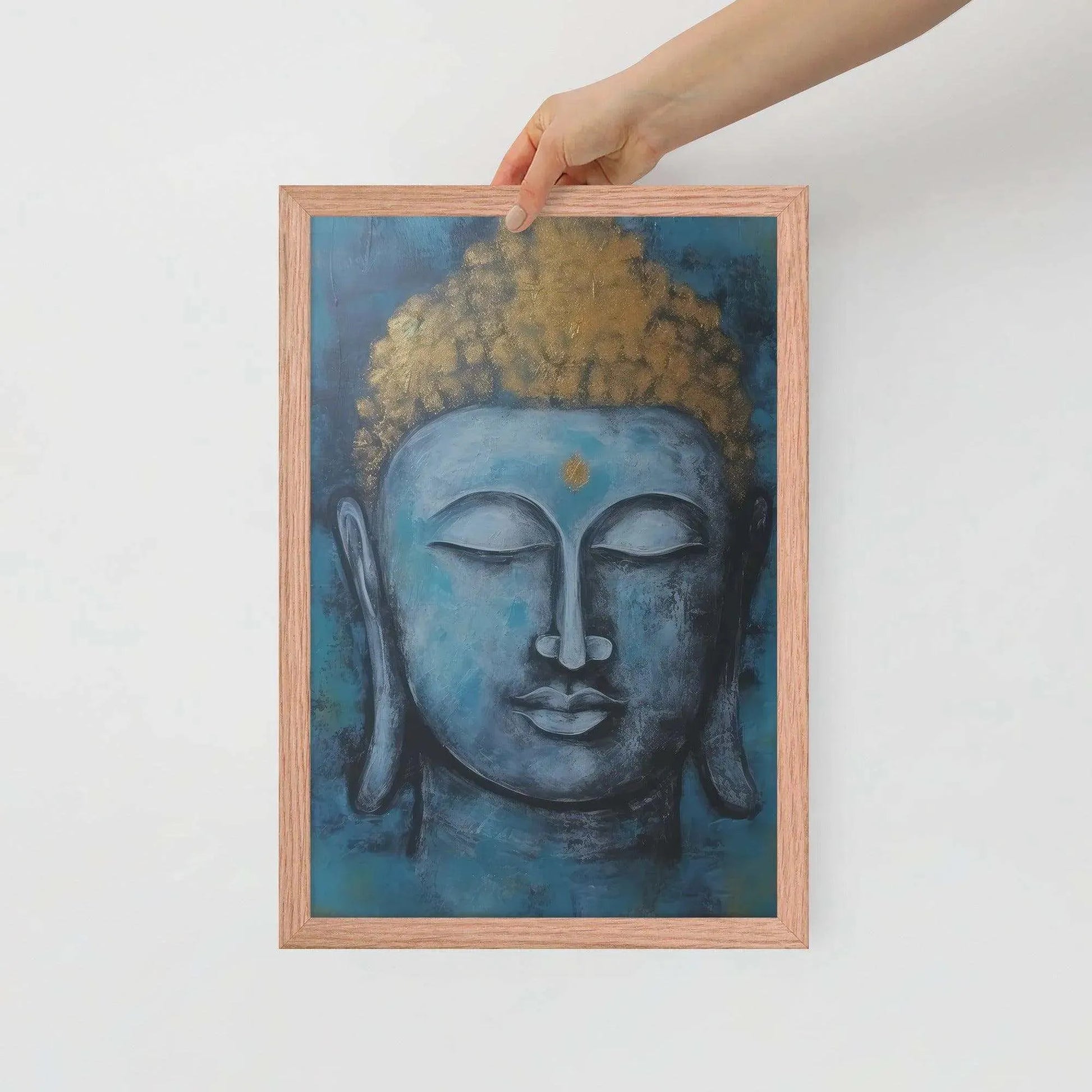 A hand is adjusting a framed poster with a red oak finish depicting a blue Buddha head with a textured gold leaf crown against a blue background, giving the impression of tranquility and artistic simplicity against a white backdrop.