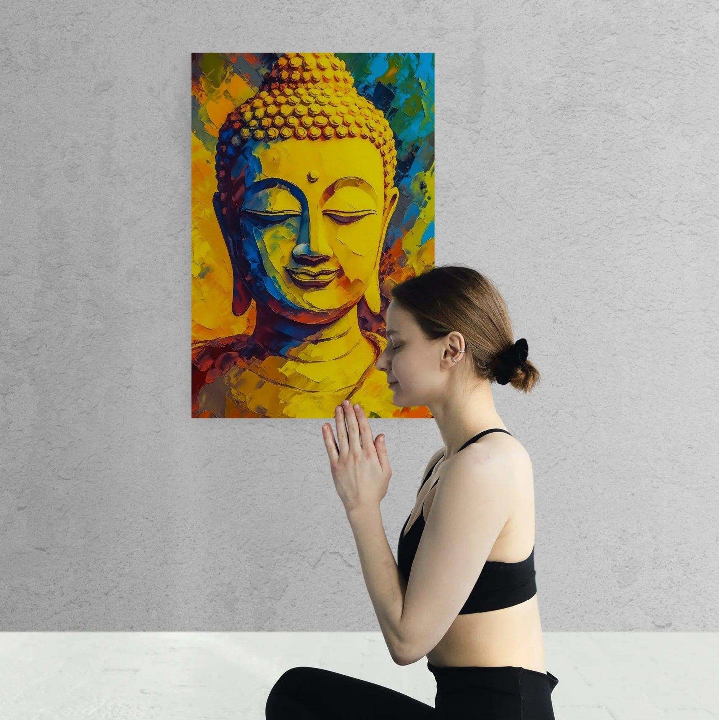 A woman in a black top performs Namaste in front of a vibrant, abstract Buddha painting with a mosaic of warm yellow, orange, and blue hues on a textured white wall.