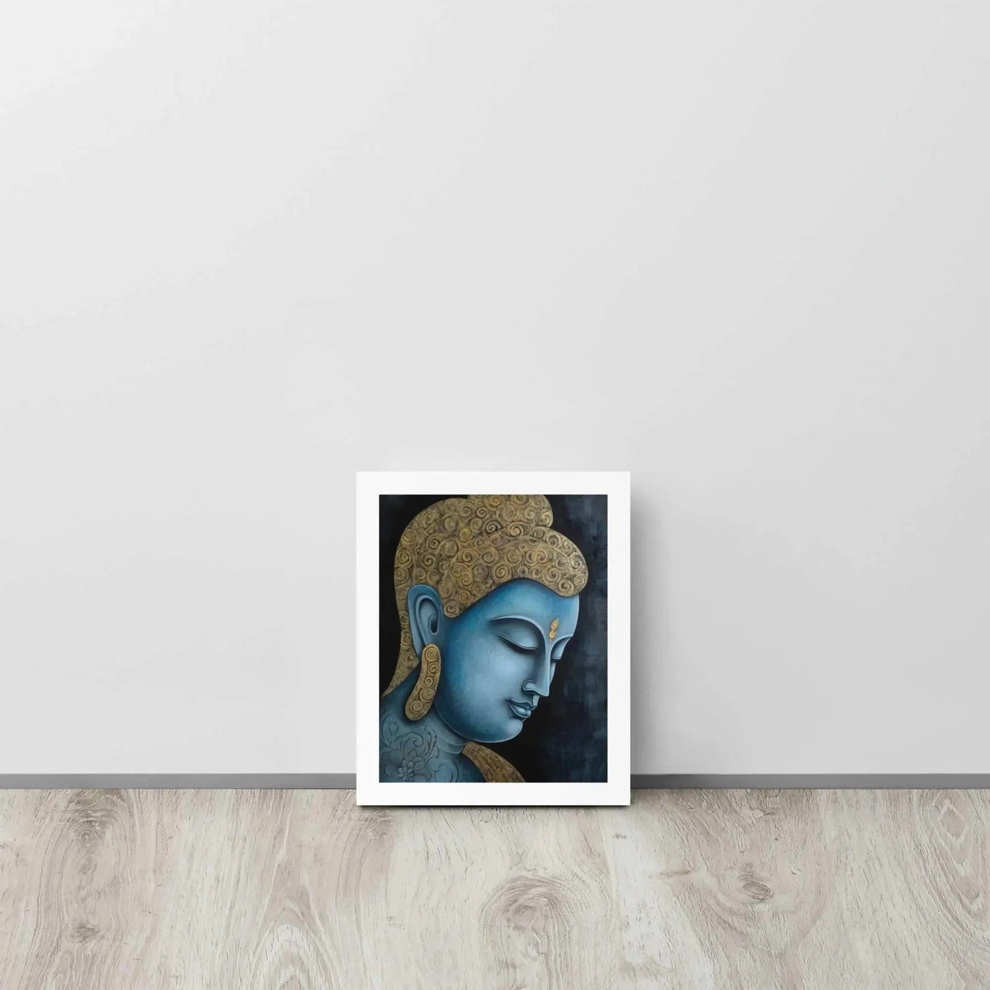 A framed Tibetan Blue Buddha Painting depicts a side profile of a serene, blue-toned Buddha with a golden patterned head, highlighted with a delicate golden ornament on the forehead. The artwork is placed against a dark, smokey background within a white oak frame, resting on a light wooden floor next to a white wall.