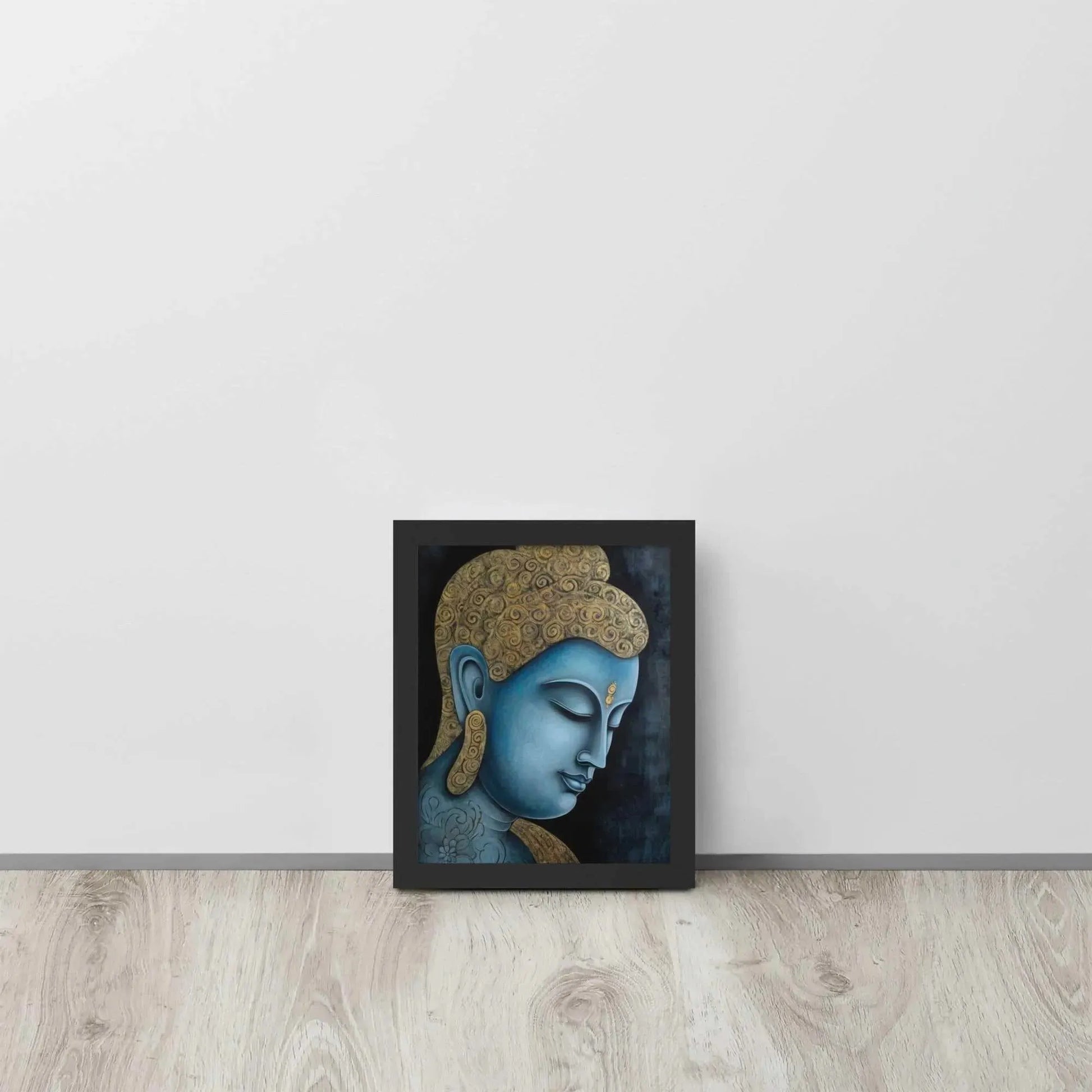 A framed Tibetan Blue Buddha Painting depicts a side profile of a serene, blue-toned Buddha with a golden patterned head, highlighted with a delicate golden ornament on the forehead. The artwork is placed against a dark, smokey background within a black oak frame, resting on a light wooden floor next to a white wall.