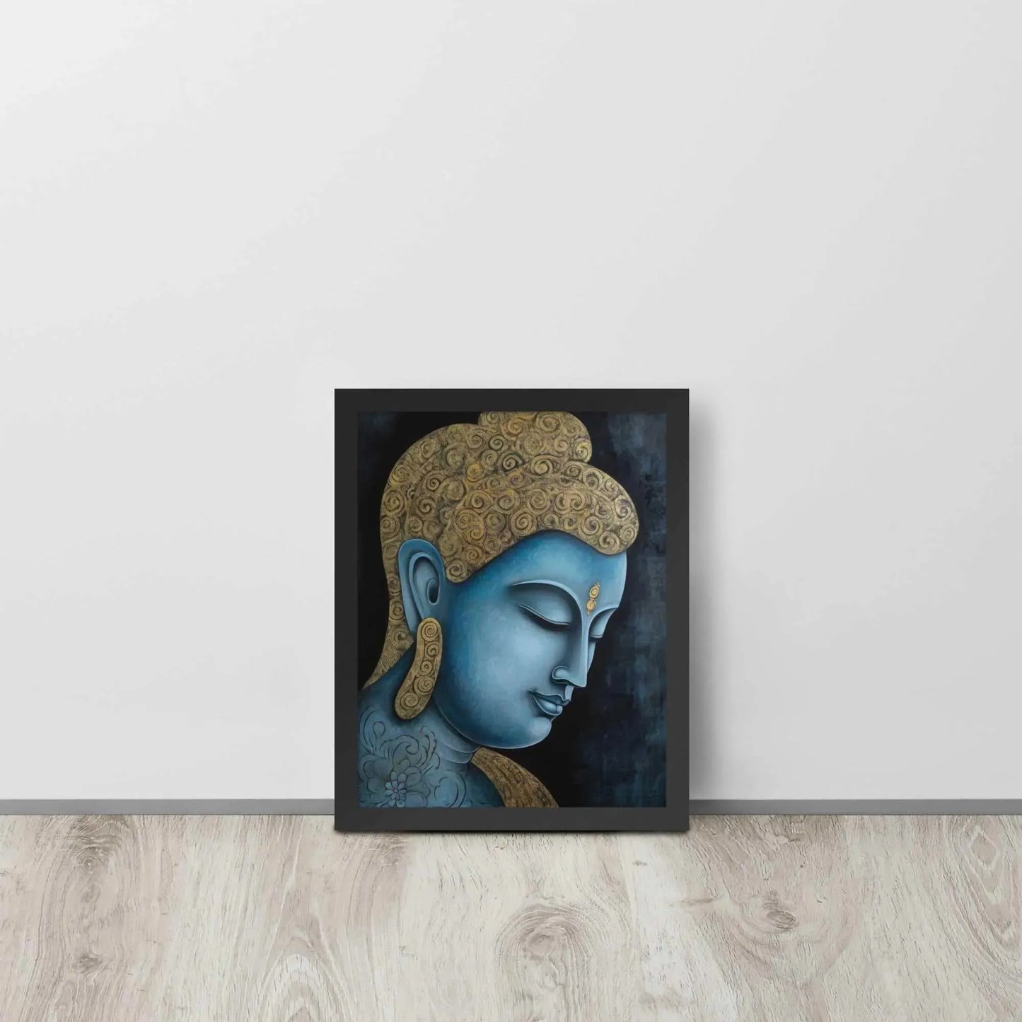 A framed Tibetan Blue Buddha Painting depicts a side profile of a serene, blue-toned Buddha with a golden patterned head, highlighted with a delicate golden ornament on the forehead. The artwork is placed against a dark, smokey background within a black oak frame, resting on a light wooden floor next to a white wall.