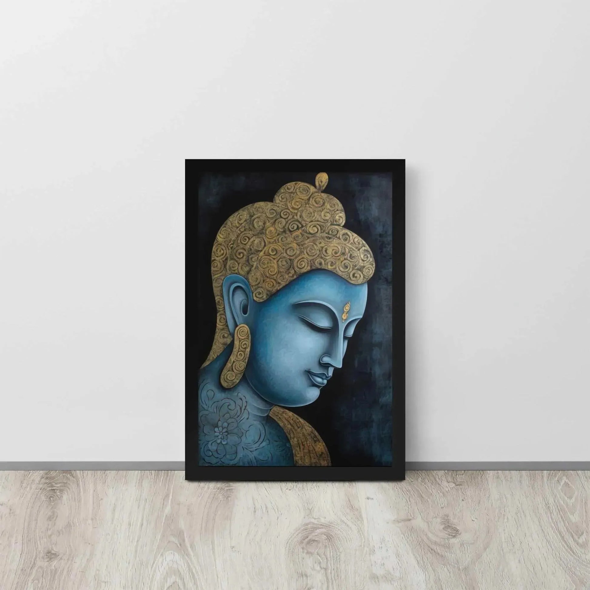 A framed Tibetan Blue Buddha Painting depicts a side profile of a serene, blue-toned Buddha with a golden patterned head, highlighted with a delicate golden ornament on the forehead. The artwork is placed against a dark, smokey background within a black oak frame, resting on a light wooden floor next to a white wall.