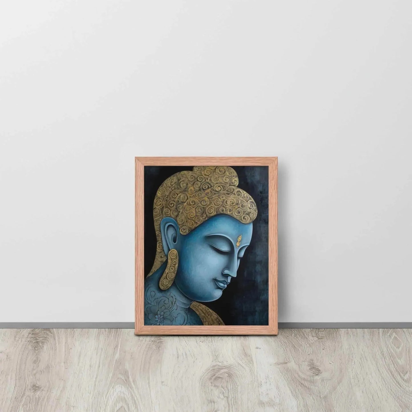 A framed Tibetan Blue Buddha Painting depicts a side profile of a serene, blue-toned Buddha with a golden patterned head, highlighted with a delicate golden ornament on the forehead. The artwork is placed against a dark, smokey background within a red oak frame, resting on a light wooden floor next to a white wall.